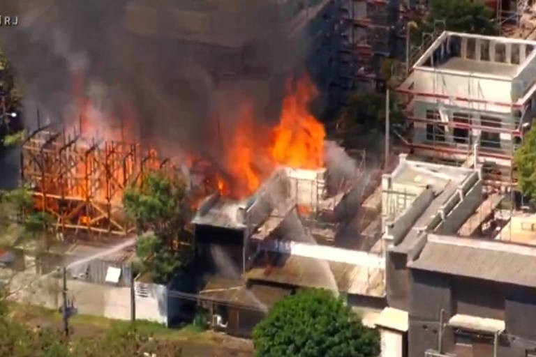 A imagem mostra um incêndio em uma construção em Curicica, com chamas e fumaça saindo de uma estrutura. Ao fundo, há várias construções em andamento e árvores ao redor. Na parte inferior da imagem, há um banner informativo com o texto 'INCÊNDIO EM CURICICA E PRAÇA SECA'.