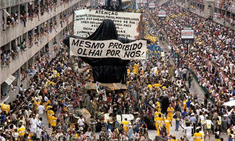A imagem mostra uma grande manifestação em uma rua movimentada, repleta de pessoas. No centro, há um carro de som coberto com um pano preto, e em cima dele, uma faixa grande com a frase 'MESMO PROIBIDO OLHAI POR NÓS!'. Ao fundo, há várias pessoas segurando cartazes e bandeiras, enquanto outras observam das janelas dos prédios ao redor. A multidão é composta por pessoas de diferentes idades e estilos, algumas usando camisetas amarelas.