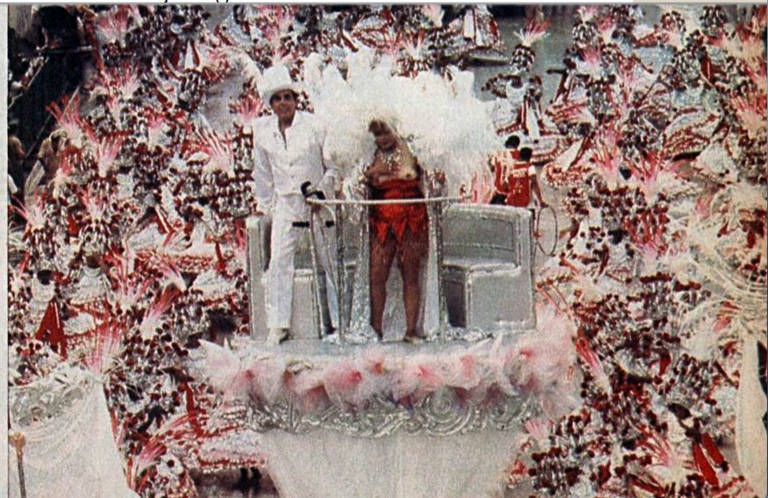 A imagem mostra um carro alegórico de carnaval, decorado com muitas flores e plumas em tons de rosa e branco. No carro, há duas pessoas: uma delas está vestida com um traje branco e a outra com um traje vermelho e plumas. Ambas estão em pé, com um fundo vibrante de decorações festivas.