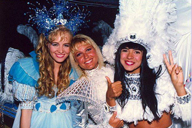 A imagem mostra três mulheres sorrindo, vestidas com trajes elaborados e coloridos. A mulher à esquerda usa um vestido azul claro com detalhes brancos e uma tiara com enfeites azuis. A mulher do meio está vestida com um traje prateado e brilhante, enquanto a mulher à direita usa um traje branco com detalhes prateados e uma pluma grande na cabeça. Todas estão em um ambiente festivo.