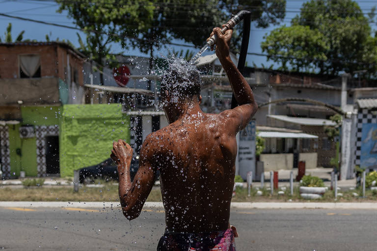 Um homem sem camisa está de costas， usando uma mangueira para se refrescar com água. Ele está em uma rua， e ao fundo há casas e árvores. A água está sendo jorrada sobre ele， criando um efeito de respingos. O céu está claro e ensolarado.