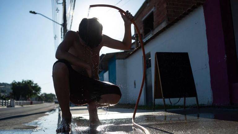 A imagem mostra uma criança agachada na calçada， segurando uma mangueira de onde sai água. A luz do sol está atrás da criança， criando um efeito de silhueta. O ambiente é urbano， com uma parede colorida ao fundo e uma placa preta visível.