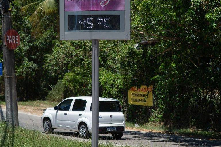 A imagem mostra um termômetro digital que indica a temperatura de 45，9°C. Ao lado， há um carro branco estacionado na estrada. No fundo， é possível ver uma placa amarela com informações de contato e uma árvore densa ao redor.