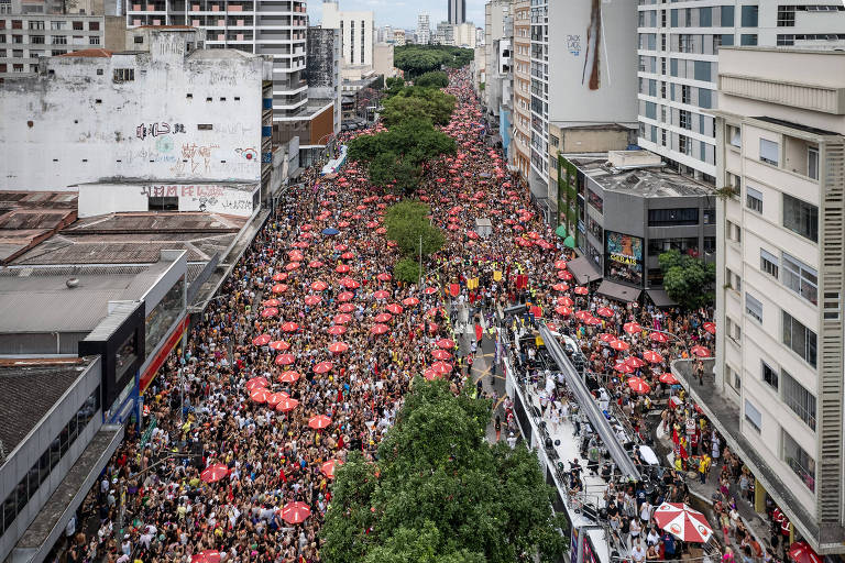 A imagem mostra uma grande multidão reunida em uma rua urbana， com pessoas usando guarda-chuvas vermelhos. O cenário é cercado por prédios altos e há uma árvore verde na parte inferior da imagem. A multidão se estende por toda a largura da rua， sugerindo um evento ou manifestação significativa.