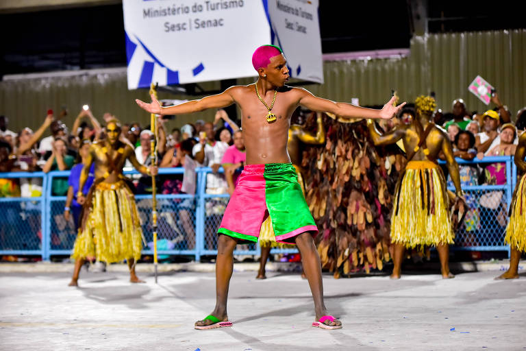 Um dançarino está se apresentando em um evento cultural. Ele é um homem sem camisa， usando um short colorido， predominantemente rosa e verde. Ao fundo， há uma multidão assistindo， com algumas pessoas vestindo roupas douradas e dançando. O ambiente parece festivo， com bandeiras e cartazes visíveis.