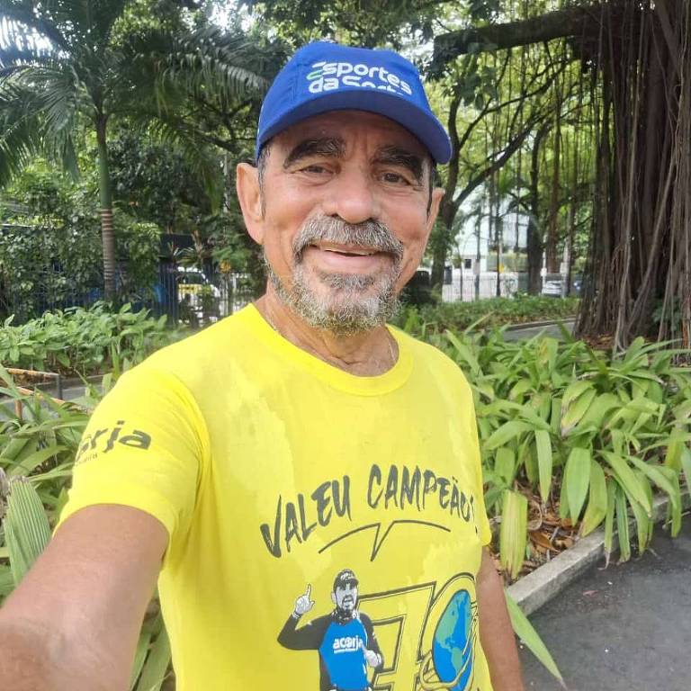 Um homem sorridente está posando para uma selfie em um ambiente ao ar livre， cercado por plantas. Ele usa uma camiseta amarela com a frase 039;Valeu Campeão039; e uma ilustração de um corredor， além de um boné azul. O fundo apresenta árvores e vegetação.