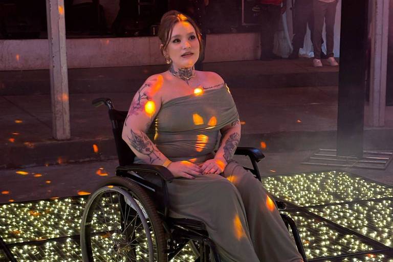 A imagem mostra uma mulher sentada em uma cadeira de rodas， vestindo um vestido longo cinza. Ela tem tatuagens visíveis nos braços e está em um ambiente com luzes brilhantes no chão， criando um efeito de festa. Ao fundo， há pessoas em pé， algumas observando， e um espaço decorado com luzes. A mulher parece estar sorrindo e aproveitando o momento.