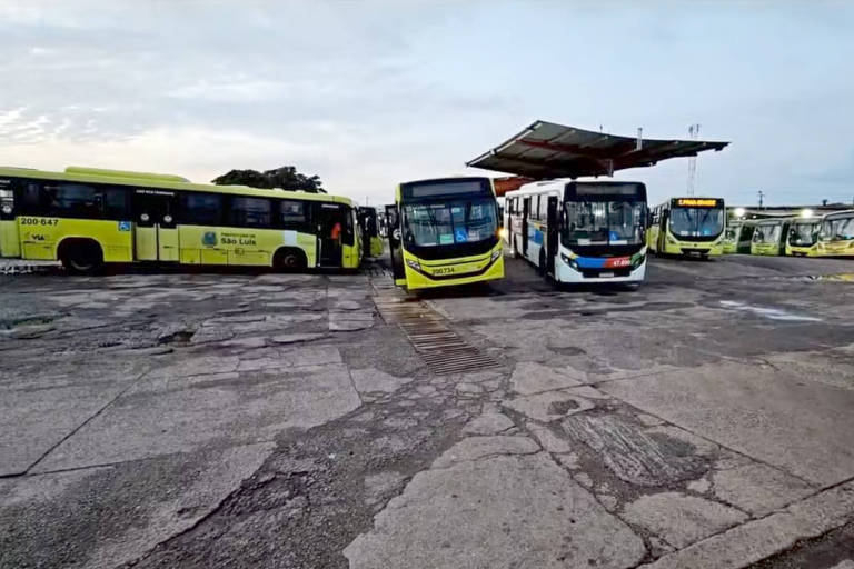 A imagem mostra uma estação de ônibus com vários veículos estacionados. Há três ônibus em destaque， sendo dois amarelos e um branco. O ambiente é ao ar livre， com um telhado coberto sobre a área de embarque. O chão é de concreto e há uma leve iluminação natural.