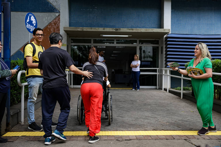 A imagem mostra um grupo de pessoas em frente a um edifício. Uma mulher em uma cadeira de rodas está sendo ajudada por um homem， enquanto outra mulher à direita está oferecendo algo. Há mais pessoas observando e um sinal azul ao fundo. O ambiente parece ser um local de atendimento ou evento comunitário.