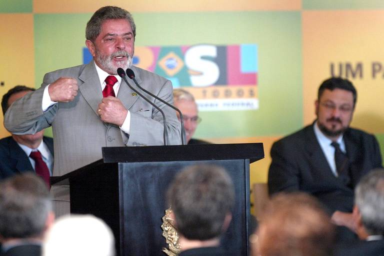 Um homem de terno cinza， com cabelo grisalho e barba， está em pé em um palco， fazendo um discurso. Ele gesticula com a mão direita， enquanto a esquerda está fechada em um punho. Ao fundo， há um painel colorido com as letras 039;BRASIL039; e 039;UM PROJETO PARA TODOS039;. Sentados à mesa， há outros homens， alguns com ternos escuros. O ambiente parece ser um evento político.