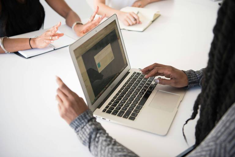 A imagem mostra uma mesa de reunião com um laptop aberto em primeiro plano. A mão de uma pessoa está digitando no teclado do laptop， enquanto outras mãos estão visíveis ao fundo， gesticulando. Há também um bloco de notas e uma caneta na mesa.