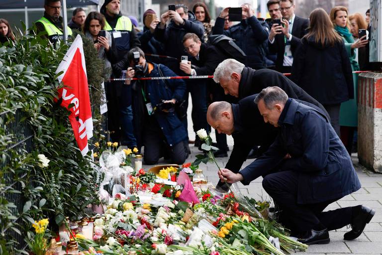 A imagem mostra um grupo de pessoas em um local de homenagem， onde há uma grande quantidade de flores e velas dispostas no chão. Duas pessoas estão se agachando para colocar flores， enquanto outras pessoas ao fundo estão tirando fotos com seus celulares. Algumas pessoas estão vestindo coletes de segurança. O ambiente parece ser urbano， com um cercado ao fundo.