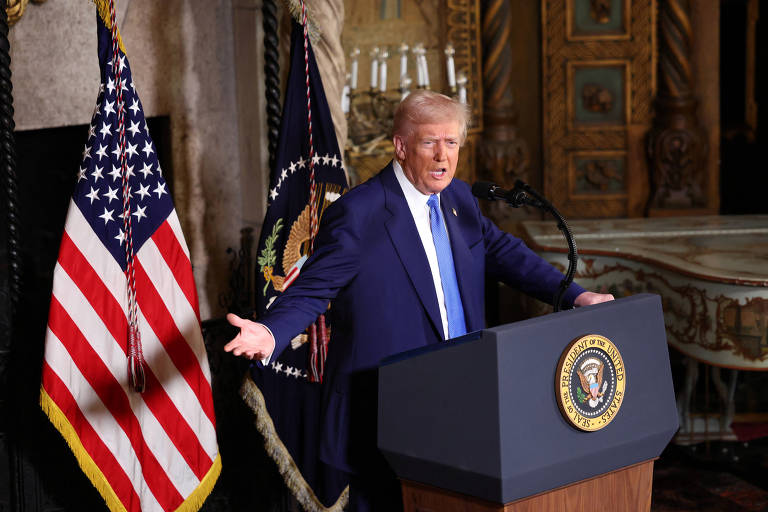 Um homem em um terno escuro e gravata azul está em pé em um palco， gesticulando com as mãos. Ao fundo， há uma bandeira dos Estados Unidos e um emblema presidencial. O ambiente é decorado de forma elaborada， com detalhes em dourado e um candelabro visível.