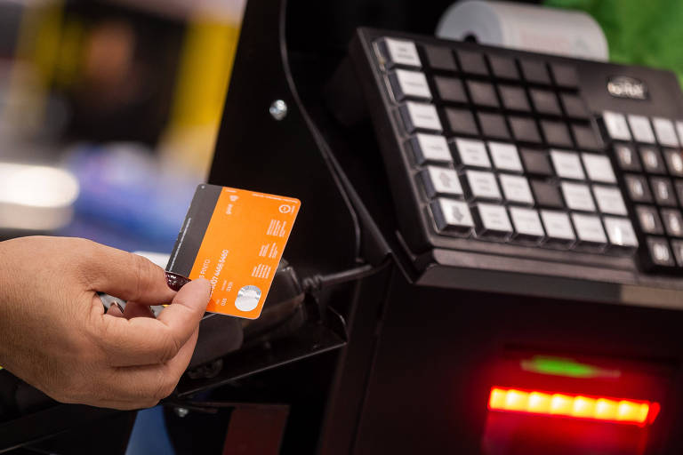 Uma mão segurando um cartão laranja próximo a um terminal de pagamento. O terminal possui um teclado com várias teclas e uma luz vermelha acesa. O fundo é desfocado, sugerindo um ambiente de loja ou mercado.