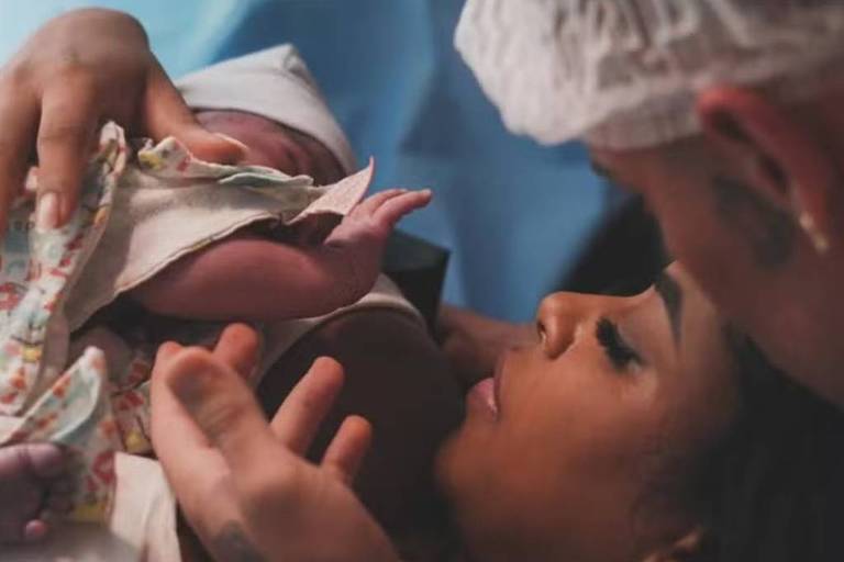 A imagem mostra uma mãe segurando seu recém-nascido, que está envolto em um cobertor. A mãe está deitada e olhando para o bebê com uma expressão de amor e admiração. Um homem, possivelmente o pai, está ao lado dela, segurando o bebê com cuidado. O ambiente parece ser uma sala de parto, com um fundo azul e elementos que sugerem um ambiente hospitalar.