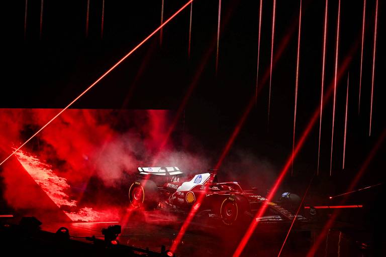 A imagem mostra um carro de Fórmula 1 em um evento de apresentação. O carro está em meio a uma atmosfera dramática, com fumaça vermelha e feixes de luz vermelha iluminando o ambiente. O fundo é escuro, destacando o carro e os efeitos visuais ao redor.