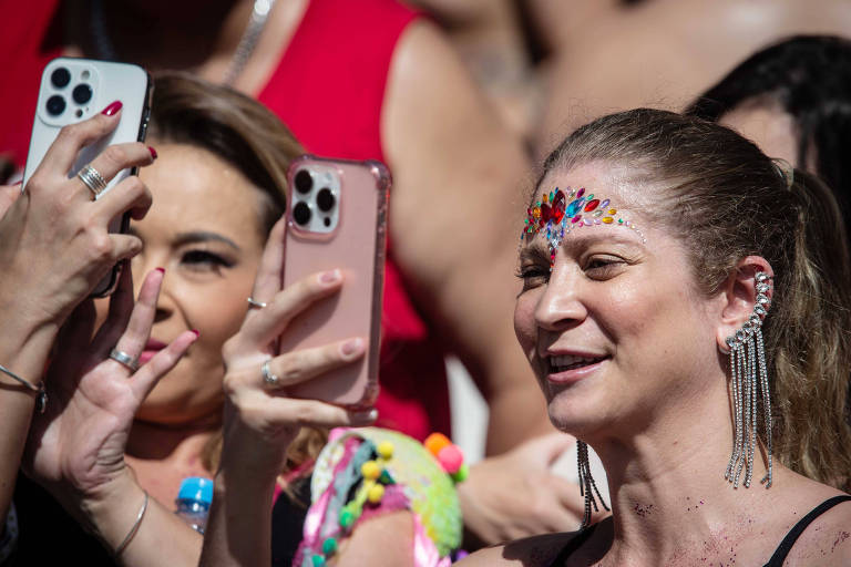 A imagem mostra um grupo de pessoas em um evento festivo， com foco em duas mulheres. A mulher à frente， com cabelo preso， usa um enfeite colorido na testa e está sorrindo enquanto segura um celular. A mulher atrás dela também está segurando um celular， com um olhar atento. Ambas parecem estar se divertindo em um ambiente animado.