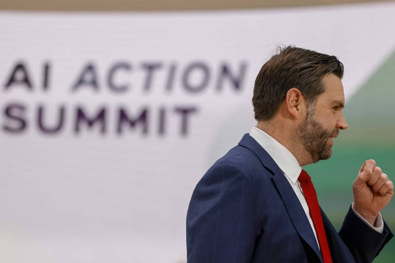 Homem de meia idade branco， de barba e cabelo escuros， aparece de perfil com o punho fechado， usando terno escuro， camisa branca e gravata vermelha， à frente de letreiro escrito: quot;AI ACTION SUMMITquot;