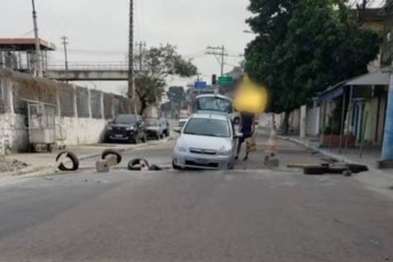 A imagem mostra uma rua urbana com um carro branco parado no centro. Ao redor， há pneus e outros obstáculos na via. No fundo， é possível ver mais veículos estacionados e uma árvore à direita. O céu está nublado， e há postes de luz ao longo da rua.