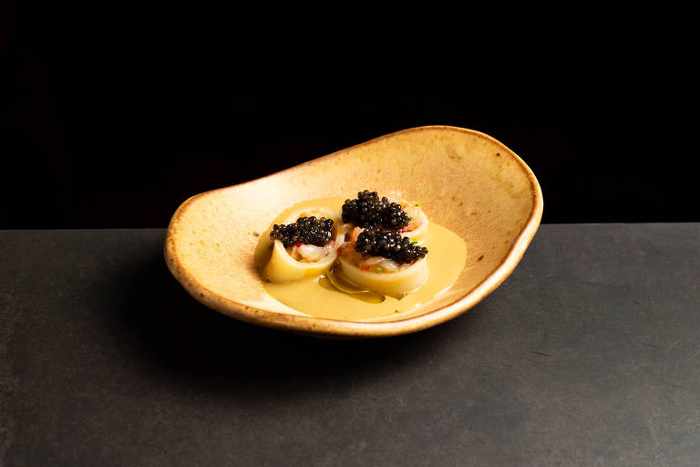 Um prato gourmet apresentado em uma tigela oval clara. O prato contém três porções de uma massa, cobertas com caviar preto, e um molho amarelo ao fundo. O fundo da imagem é escuro, destacando o prato.