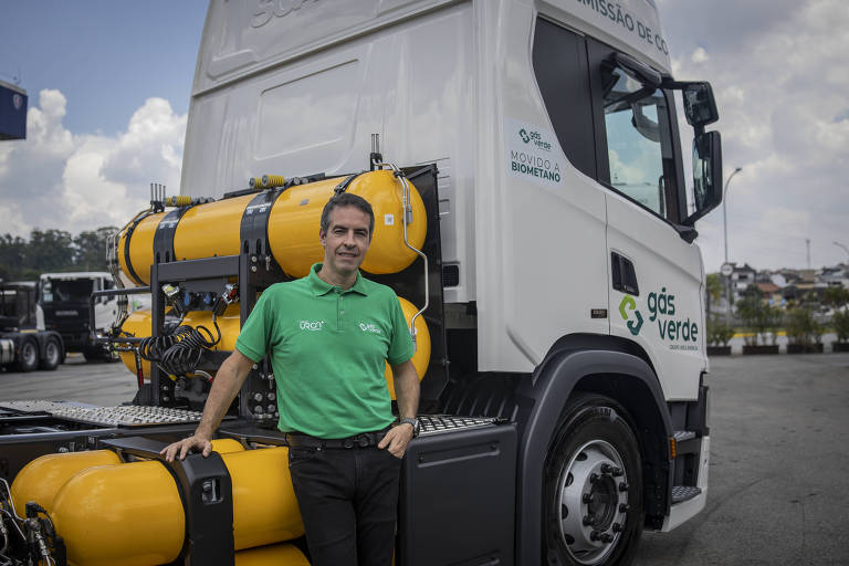 Um homem em uma camiseta verde está posicionado ao lado de um caminhão branco. O caminhão possui tanques amarelos de gás verde na parte traseira e apresenta o logotipo 'gás verde' em um dos lados. O fundo mostra um céu parcialmente nublado e uma área urbana ao longe.