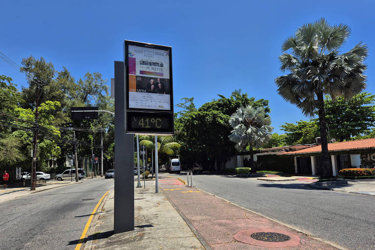 A imagem mostra um ponto de ônibus em uma rua arborizada. Há um painel publicitário com informações visíveis e a temperatura de 41°C. O céu está claro e azul， e há várias árvores ao redor， além de algumas palmeiras. No fundo， é possível ver um veículo e casas ao longo da rua.