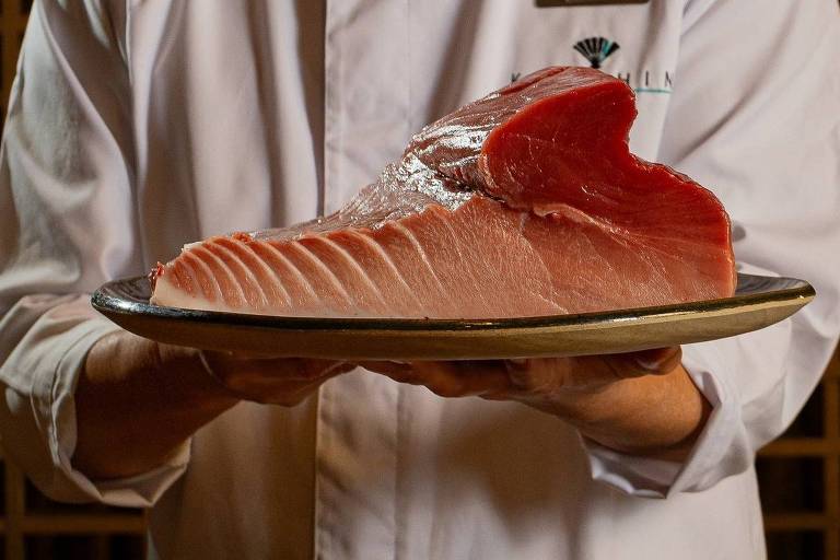 Um chef está segurando um prato com uma grande peça de atum fresco. O atum tem uma cor rosa vibrante e está bem apresentado. O chef está usando um avental branco e a imagem tem um fundo de madeira que sugere um ambiente de restaurante.