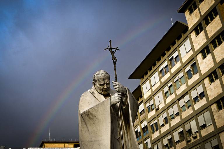 A imagem mostra uma estátua de um papa, que está de pé com uma cruz em uma das mãos e a cabeça inclinada em um gesto de oração. Ao fundo, há um céu nublado com um arco-íris visível. O ambiente ao redor é urbano, com um edifício moderno à direita da imagem.