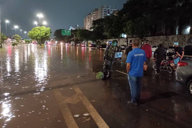 A imagem mostra uma rua inundada durante a noite， com água cobrindo a pista. Há várias pessoas na calçada， algumas observando a situação. Ao fundo， é possível ver veículos parados e árvores iluminadas por postes de luz. O céu está escuro， indicando que é de noite.