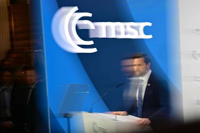 A imagem mostra Vance em um palco, aparentemente fazendo uma apresentação. Ele está em frente a um microfone e um suporte para documentos. O fundo é azul com o logotipo da MSC visível. A imagem está ligeiramente desfocada, o que dá a impressão de movimento.