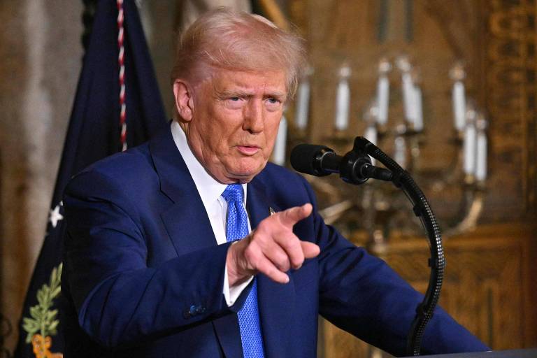 Um homem com cabelo loiro e terno azul está falando em um microfone. Ele aponta com a mão direita enquanto se dirige a uma audiência. Ao fundo， há uma bandeira e candelabros.