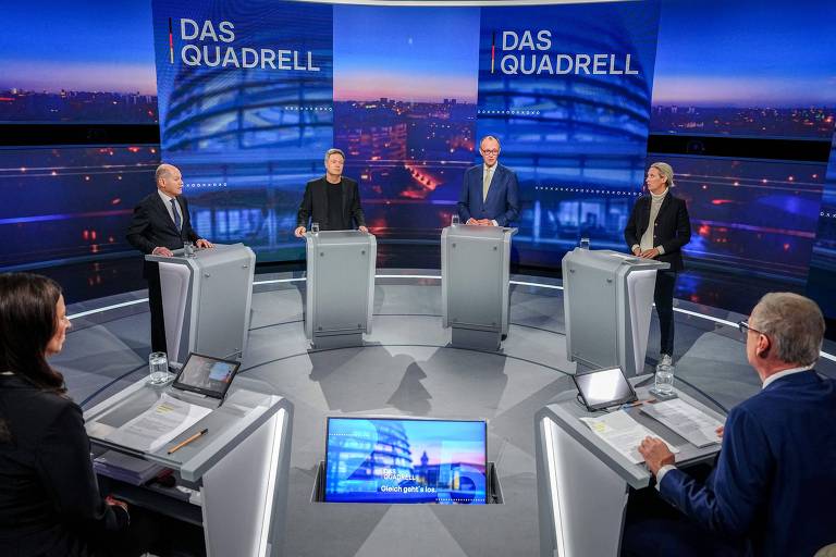 A imagem mostra um estúdio de debate com quatro participantes em pé atrás de mesas. O fundo é iluminado com uma vista da cidade ao entardecer. Os participantes estão em trajes formais， e há equipamentos de gravação visíveis. Na mesa à frente， há papéis e copos de água.