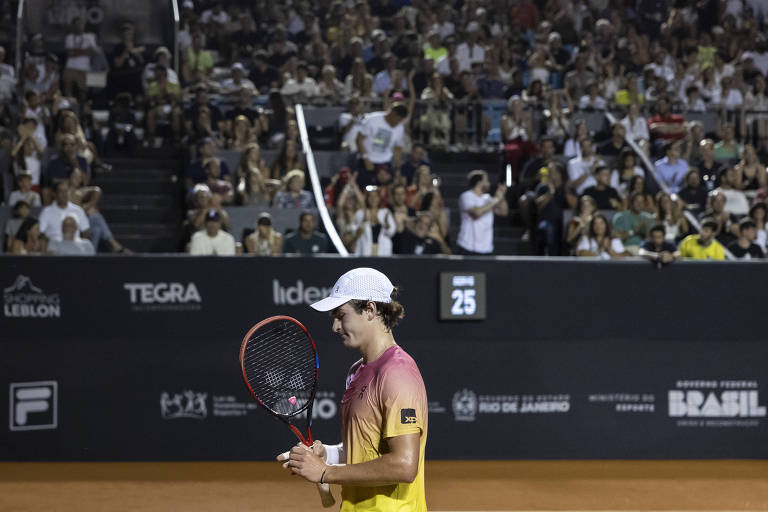 Um jogador de tênis está em quadra， segurando uma raquete. Ele usa uma camiseta com gradiente de cores que vai do rosa ao amarelo e um boné branco. Ao fundo， uma grande plateia observa atentamente， com várias pessoas visíveis nas arquibancadas. O placar ao fundo mostra o número 25. O ambiente é iluminado， sugerindo que o jogo está acontecendo à noite.