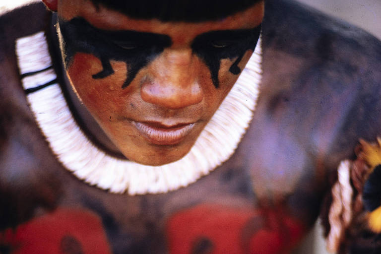 Um homem indígena está sentado， com a cabeça baixa， concentrado em suas mãos. Ele possui pintura corporal em seu torso， com desenhos em preto e vermelho， incluindo figuras que lembram rostos. O homem usa um colar de penas brancas e tem adornos coloridos em seus braços. Ele segura um objeto colorido， possivelmente um leque ou um acessório feito de penas.