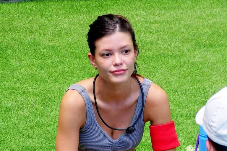  A imagem mostra uma mulher sentada em um gramado verde. Ela usa uma blusa cinza com listras e tem um acessório vermelho no braço. A mulher parece estar em uma conversa, com uma expressão pensativa. Ao fundo, é possível ver pessoas, mas não estão claramente visíveis.
