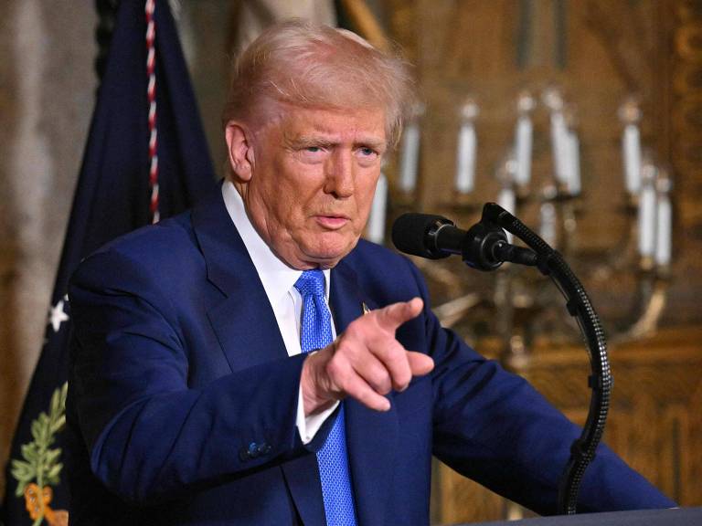 Um homem com cabelo loiro e terno azul está falando em um microfone. Ele aponta com a mão direita enquanto se dirige a uma audiência. Ao fundo， há uma bandeira e candelabros.