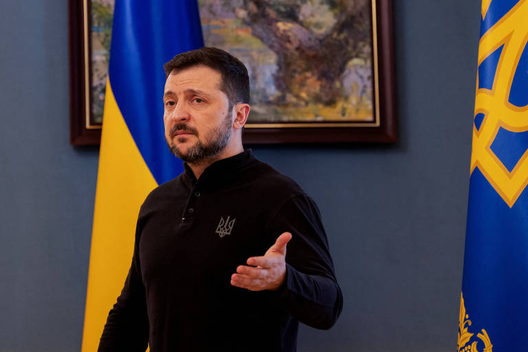 Um homem com cabelo curto e barba， vestido com uma camisa preta， está em pé em um ambiente interno. Ao fundo， há uma bandeira da Ucrânia em azul e amarelo e uma pintura emoldurada. O homem parece estar gesticulando enquanto fala.