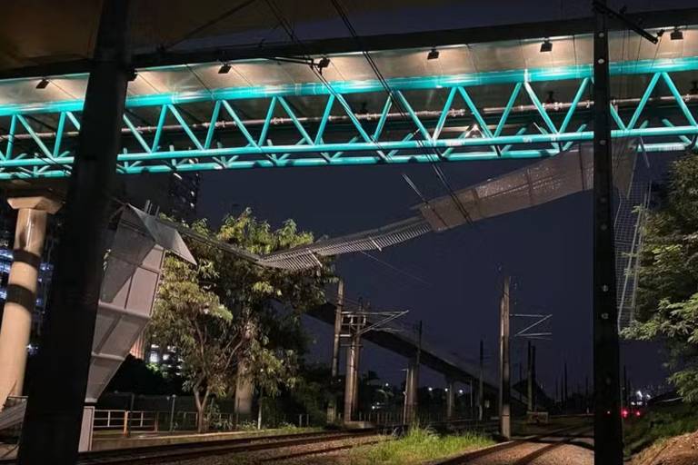 A imagem mostra uma estrutura de ponte elevada com iluminação em tom verde， vista à noite. A ponte parece estar em um estado de desvio ou colapso em uma parte， com seções inclinadas. Abaixo， há trilhos de trem e vegetação ao redor， com árvores visíveis ao fundo.