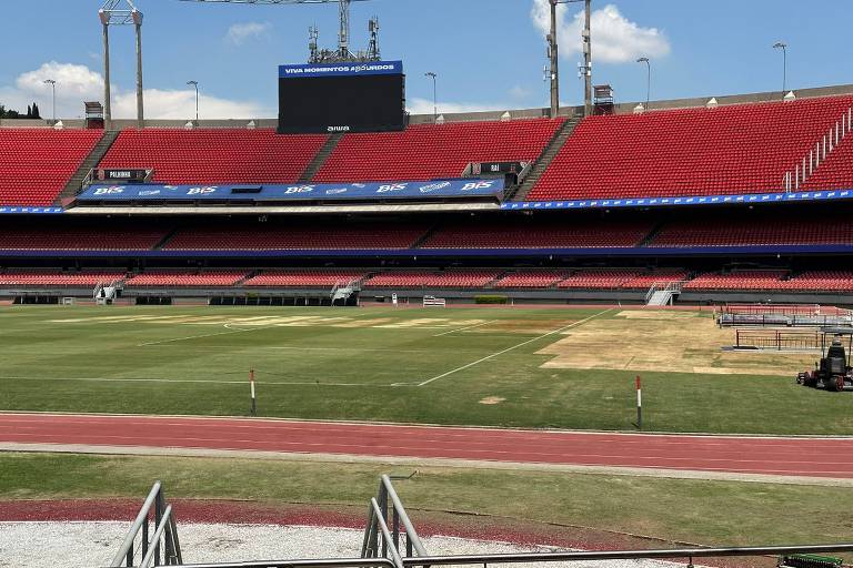 Imagem de um estádio de futebol com arquibancadas vermelhas e um campo visivelmente dividido entre áreas verdes e secas. No fundo， há um telão e a iluminação do estádio. O campo apresenta algumas marcas de manutenção， e há uma pista de atletismo ao redor do campo.