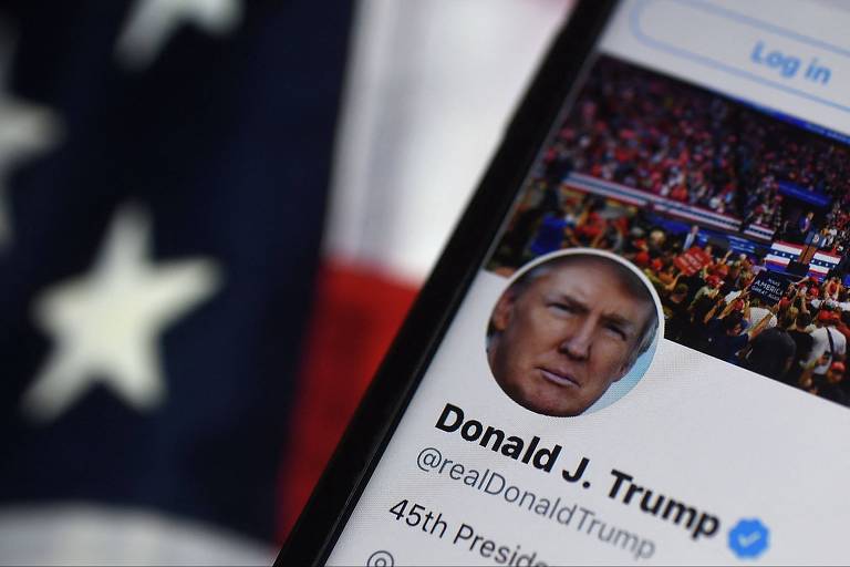 A imagem mostra a tela de um smartphone exibindo o perfil do Twitter de Donald J. Trump， com uma foto de perfil dele e um fundo de uma multidão em um evento. Ao fundo， há uma bandeira dos Estados Unidos， com listras vermelhas e brancas e estrelas brancas em um campo azul.