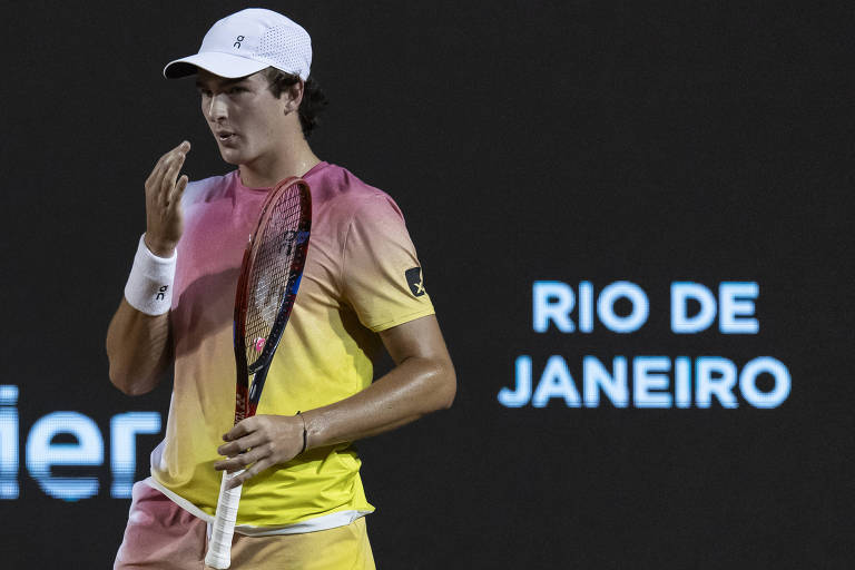 Um jogador de tênis está em uma quadra， usando uma camiseta com gradiente de cores rosa e amarelo， e shorts claros. Ele segura uma raquete e parece estar se preparando para um ponto， com uma expressão concentrada. Ao fundo， há um letreiro que diz 039;RIO DE JANEIRO039;.