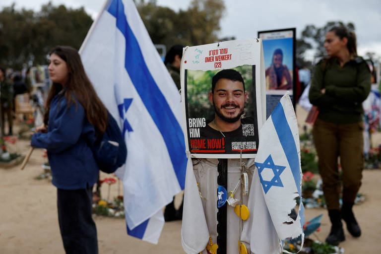 A imagem mostra um memorial com bandeiras de Israel e um cartaz que diz 039;BRING HIM HOME NOW!039; com a foto de um homem sorridente. Ao fundo， há pessoas em um ambiente ao ar livre， algumas segurando bandeiras. O cenário parece ser um local de homenagem ou protesto.