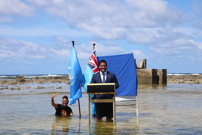 Um homem em traje formal está em pé atrás de um púlpito， falando em uma conferência， enquanto outro homem está parcialmente submerso na água ao lado dele. Ao fundo， há bandeiras da ONU e de um país， além de uma estrutura de concreto. O céu está parcialmente nublado e o mar é visível ao fundo.