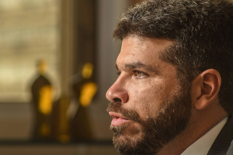 A imagem mostra um homem com barba e cabelo escuro， em perfil. Ele está em um ambiente interno， com luz natural entrando pela janela ao fundo. No fundo， há algumas figuras decorativas em tons dourados. O homem parece estar pensativo.