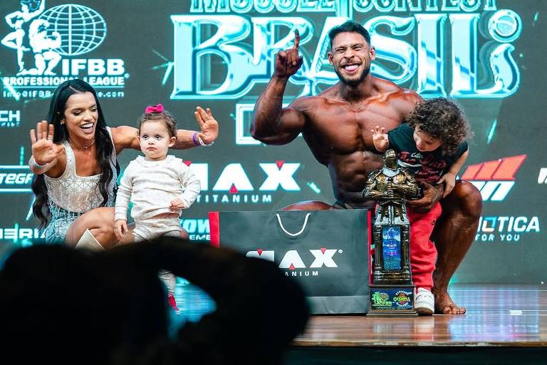 Ainda no palco， fisiculturista campeão posa para foto junto de sua esposa e seus filhos