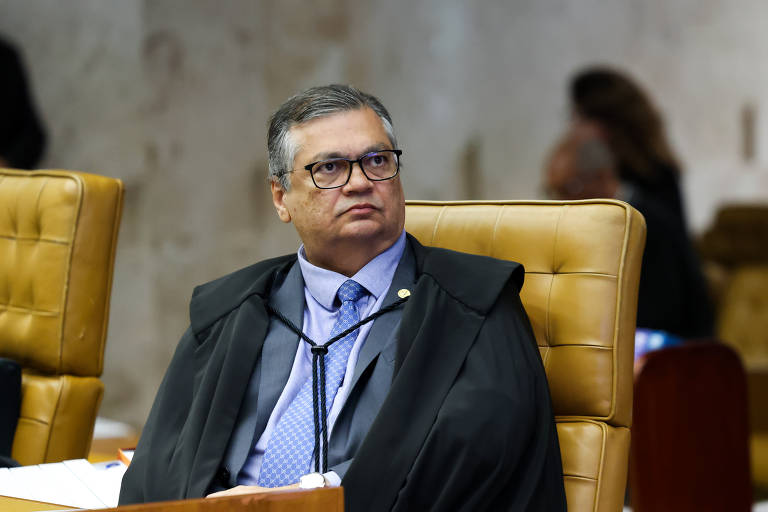 Um juiz sentado em uma cadeira de tribunal， usando um manto preto e óculos. Ele parece estar ouvindo atentamente， com uma expressão séria. O fundo é desfocado， mostrando outras pessoas no tribunal.