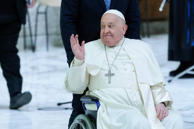 Imagem do Papa Francisco sentado em uma cadeira de rodas， acenando com a mão. Ele está vestido com uma túnica branca e um cordão com um crucifixo. Ao fundo， há pessoas e um ambiente interno com piso de mármore.