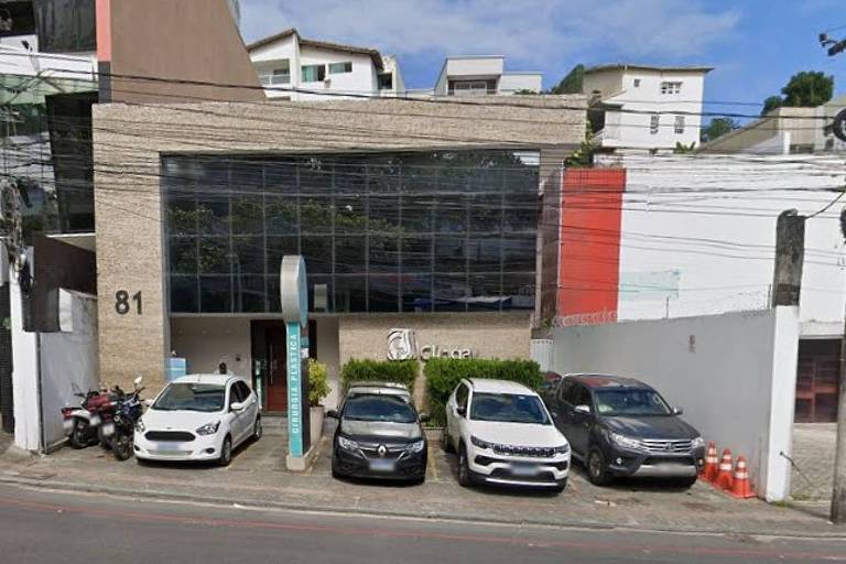 A imagem mostra a fachada de um edifício comercial de dois andares， com uma grande área de vidro na frente. O número 81 está visível na parede. Na calçada， há vários carros estacionados， incluindo um carro branco， um carro preto， um SUV e uma moto. O ambiente é urbano， com algumas árvores e um céu parcialmente nublado ao fundo.