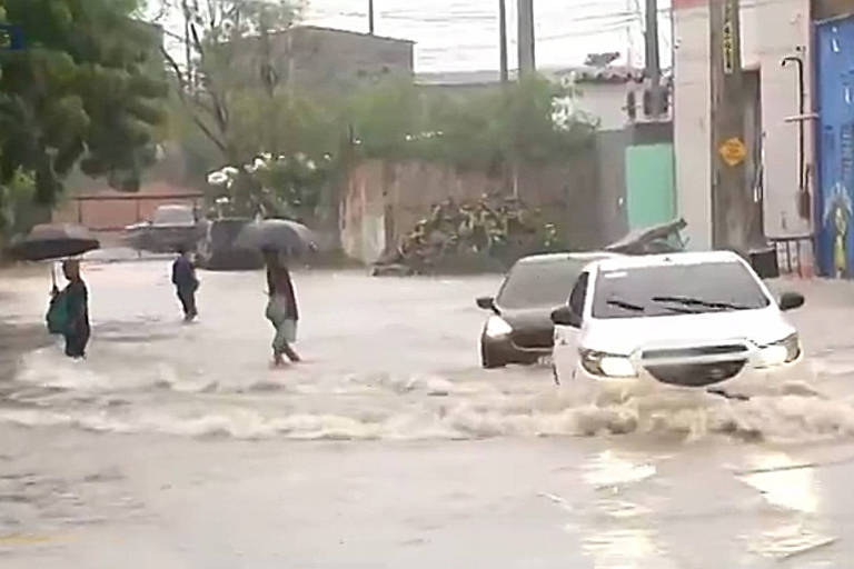 A imagem mostra uma cena de inundação em uma área urbana， com ruas alagadas. Algumas pessoas estão caminhando com guarda-chuvas， enquanto um carro branco tenta passar pela água. Ao fundo， há prédios e vegetação， e a água parece estar em um nível elevado， dificultando a passagem de veículos.