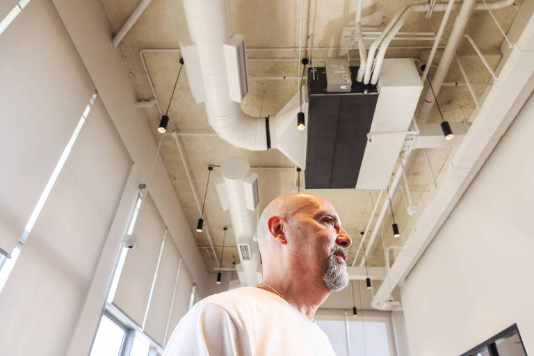 A imagem mostra um homem de perfil em um ambiente interno moderno. O teto é alto e possui dutos de ar e iluminação pendente. As paredes têm janelas grandes com cortinas. O homem tem cabelo curto e barba， vestindo uma camiseta clara.